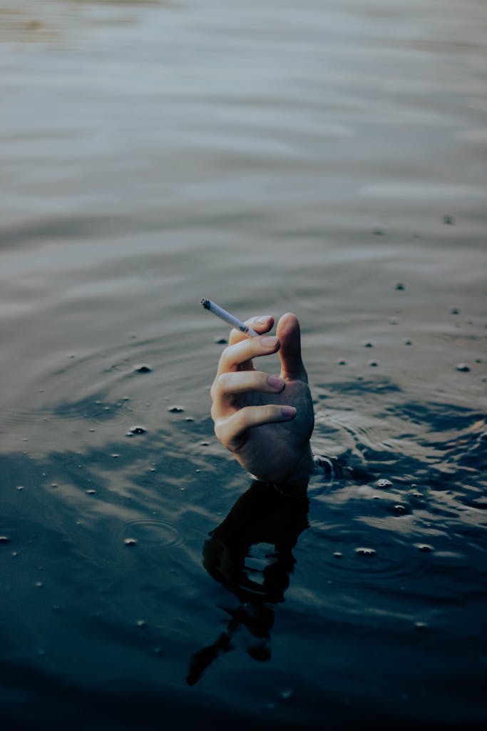 A hand emerging from water holds a cigarette, symbolizing addiction and survival.