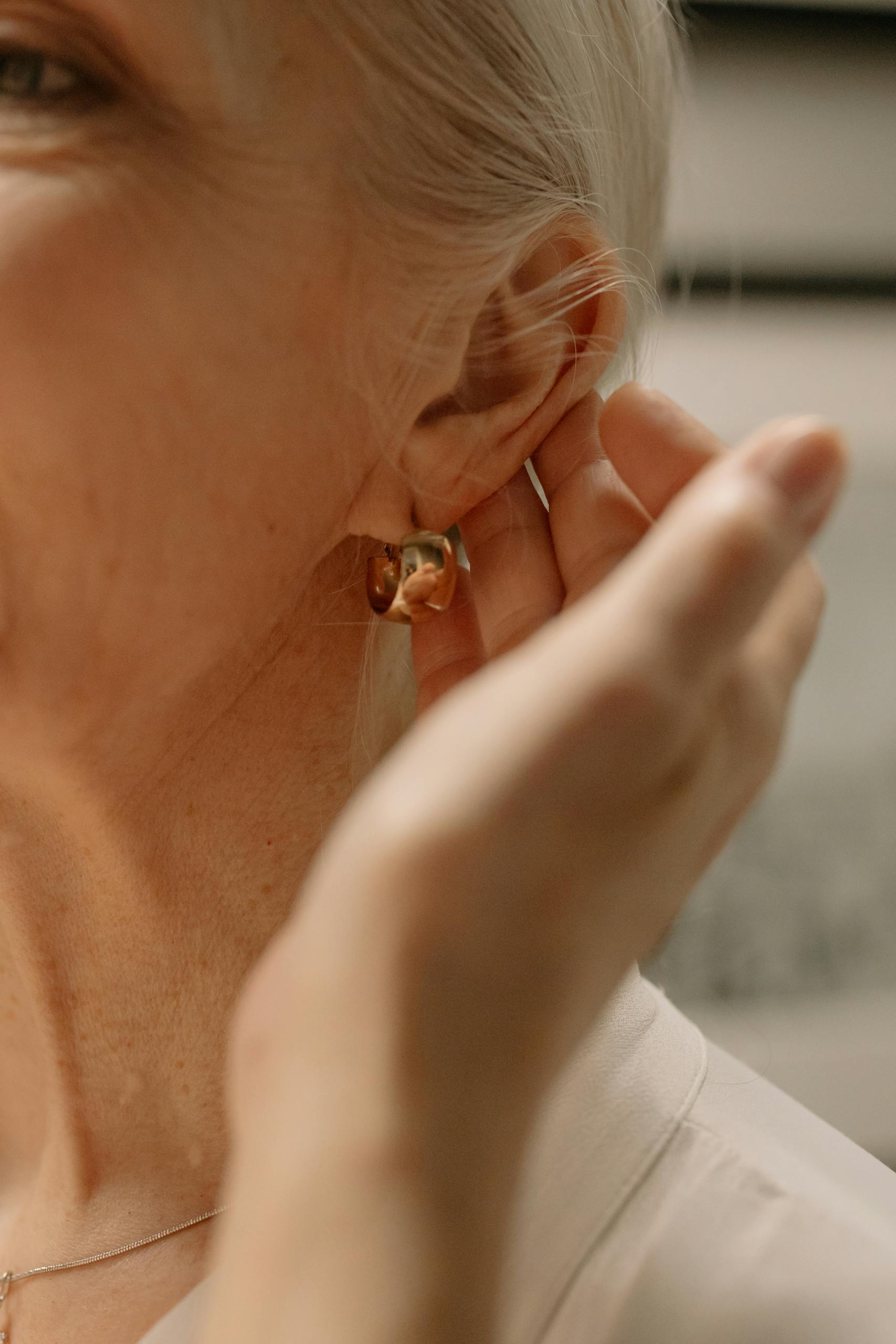 Close-up of a senior woman adjusting her stylish gold hoop earring with a soft focus background.