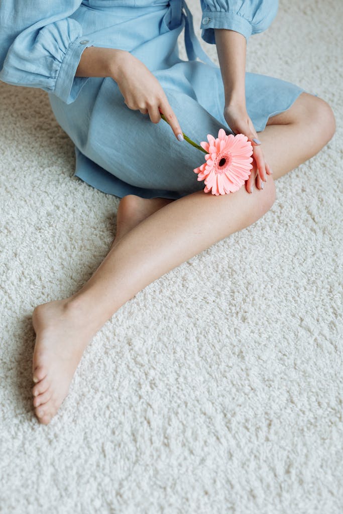 A woman in a blue dress sitting on a carpet, holding a pink gerbera daisy.