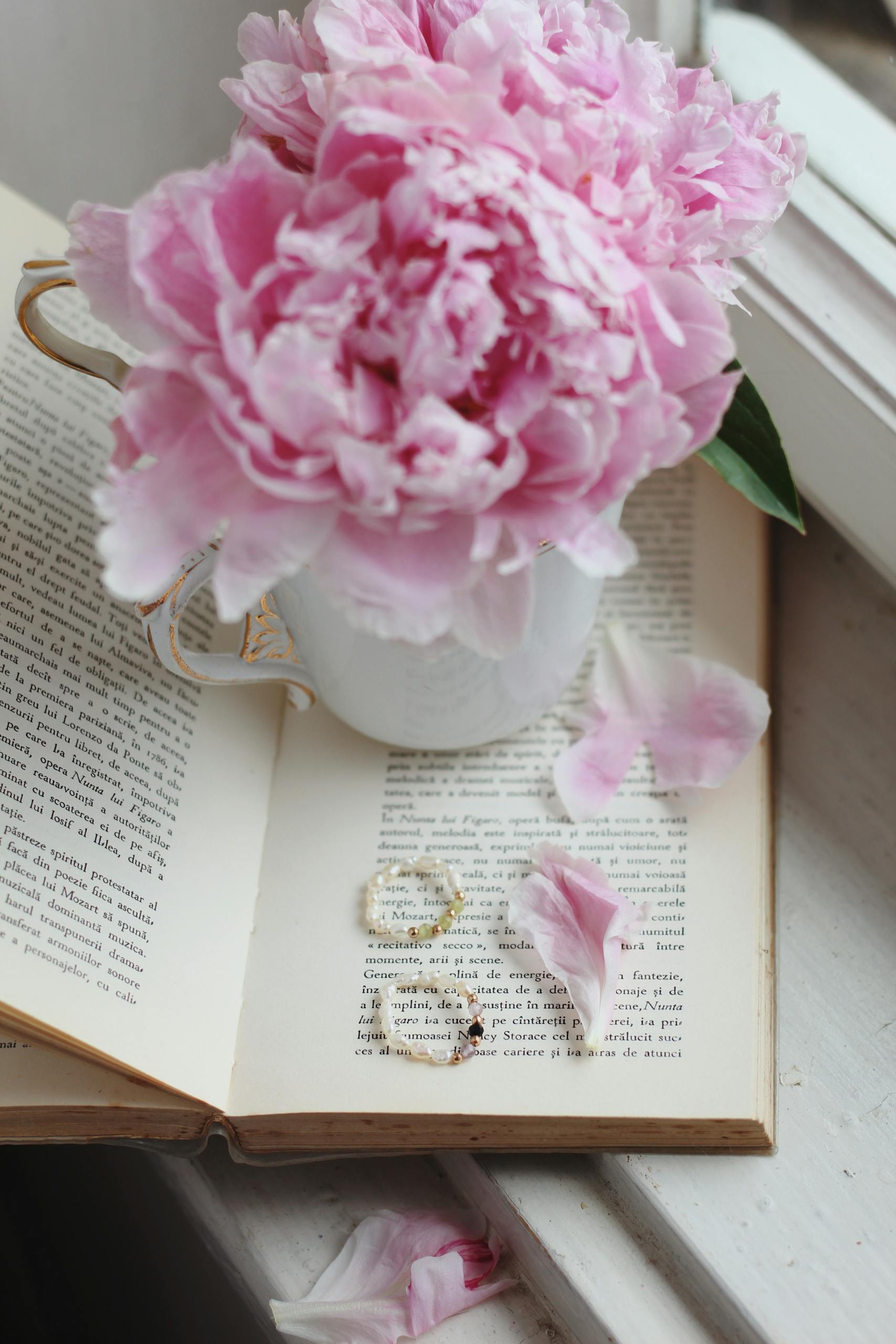 Charming pink peonies in a teacup placed on an open book, with delicate jewelry nearby, perfect for aesthetics.