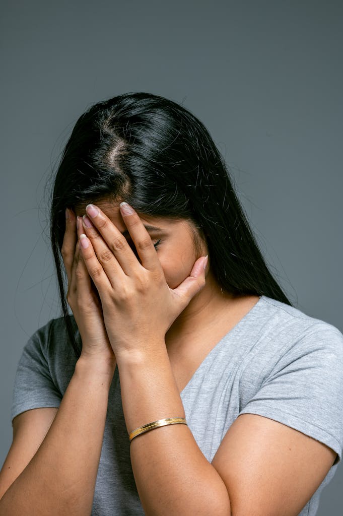 A woman in a gray shirt hides her face in hands expressing stress and emotion.
