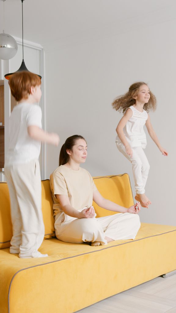 A serene woman meditates on a sofa while two children energetically jump beside her.